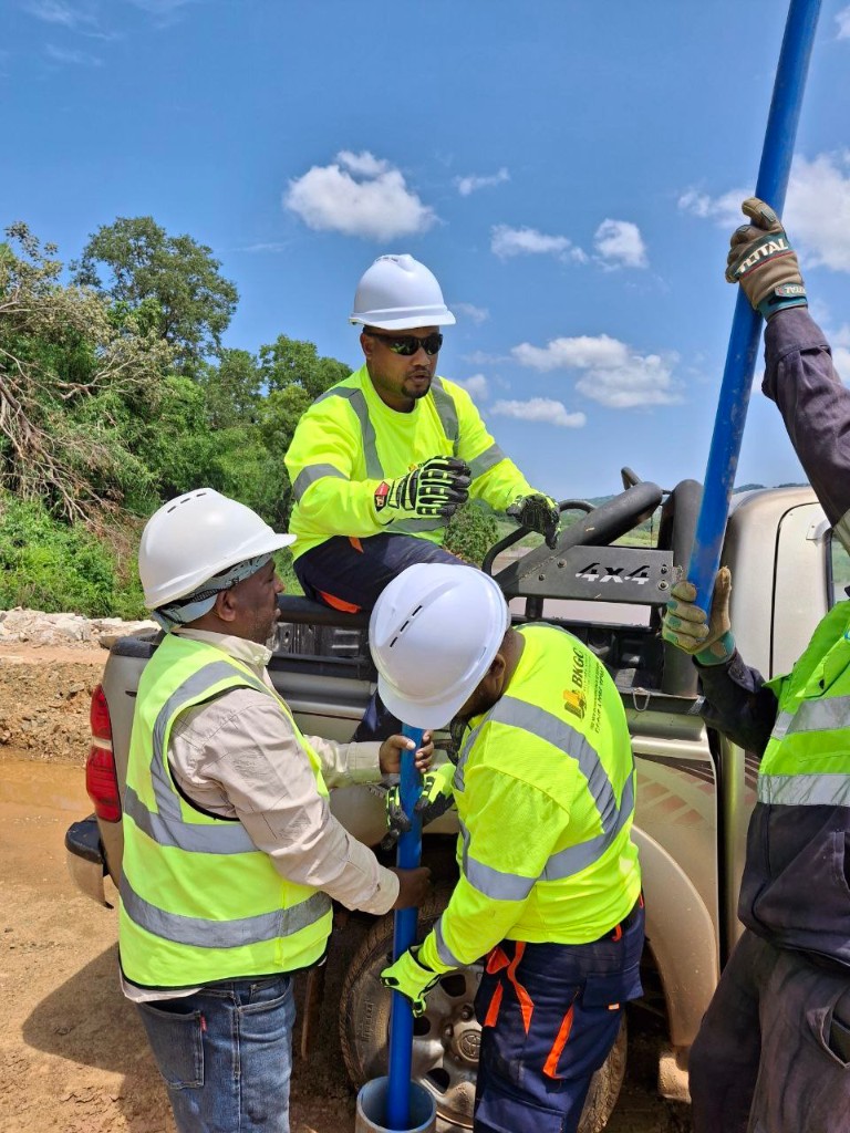 A field crew in hard hats lowering a blue well casing beside a pickup truck